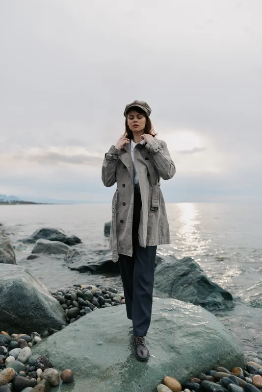 Young woman in warm coat and hat stands on rocky shore near calm sea under cloudy sky. Fashionable autumn outfit with neutral tones creates moody and contemplative outdoor scene.