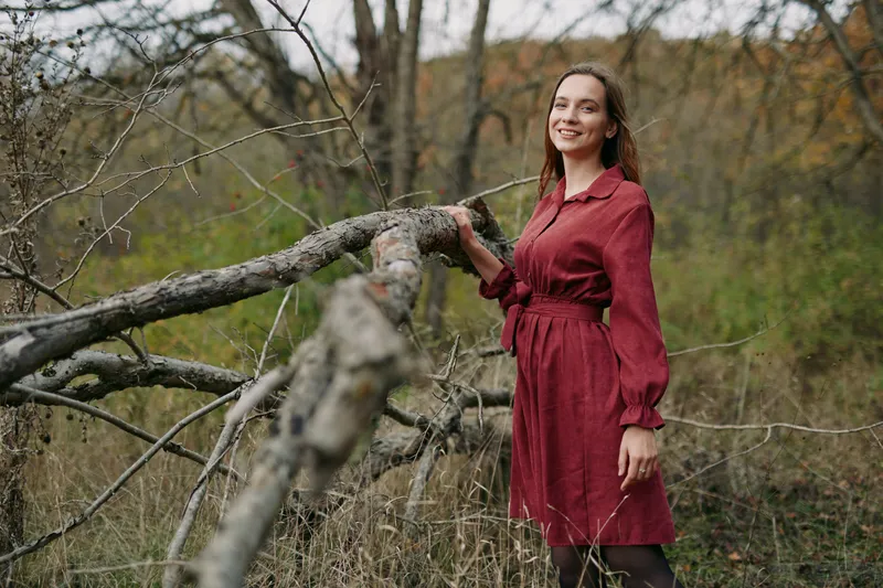 A young woman in a red dress stands beside a fallen tree in a quiet woodland, captured with soft natural light and a genuine smile to create a believable, intimate everyday portrait.