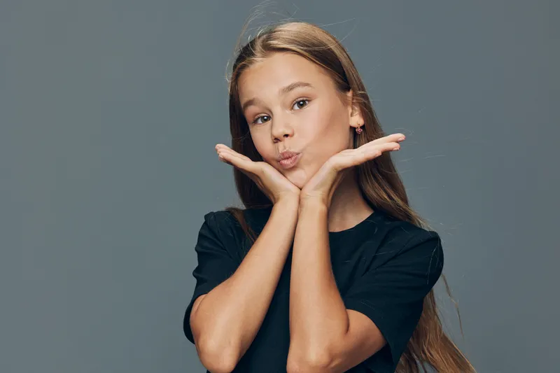 Girl with long hair and black t shirt making playful face gesture with hands under chin on gray background. Youthful expression and casual style in studio shot.
