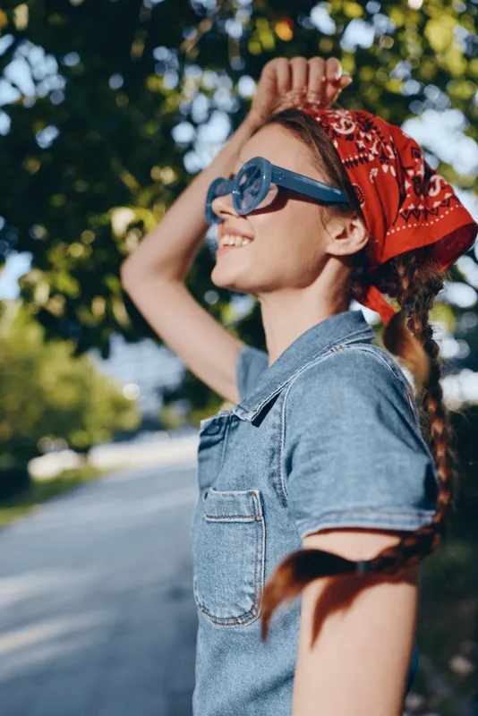Lifestyle woman in modern boho-western denim street style wearing sunglasses and a red bandana, enjoying sunny outdoor moment with film color effect and casual summer vibe.