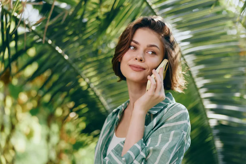 Portrait of a woman talking on a smartphone in a sunny tropical garden with palm leaves, relaxed and cheerful.