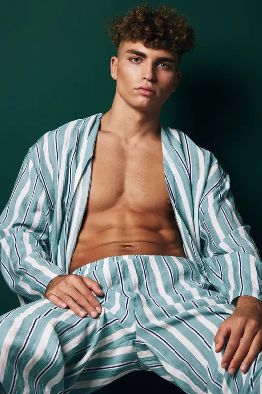 Young man with curly hair and serious face wearing striped outfit sitting and posing in studio with dark background. Fashionable male model displaying toned body with open shirt and relaxed pants.