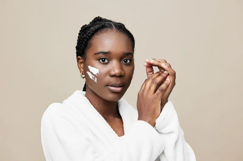 Young African woman applying facial cream, showcasing natural beauty and skincare routine, calm expression, minimalistic background, soft tones for beauty concepts
