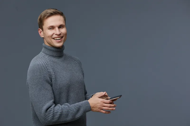 Cheerful young man in gray turtleneck sweater using smartphone smiling friendly portrait indoor studio shot. People lifestyle concept