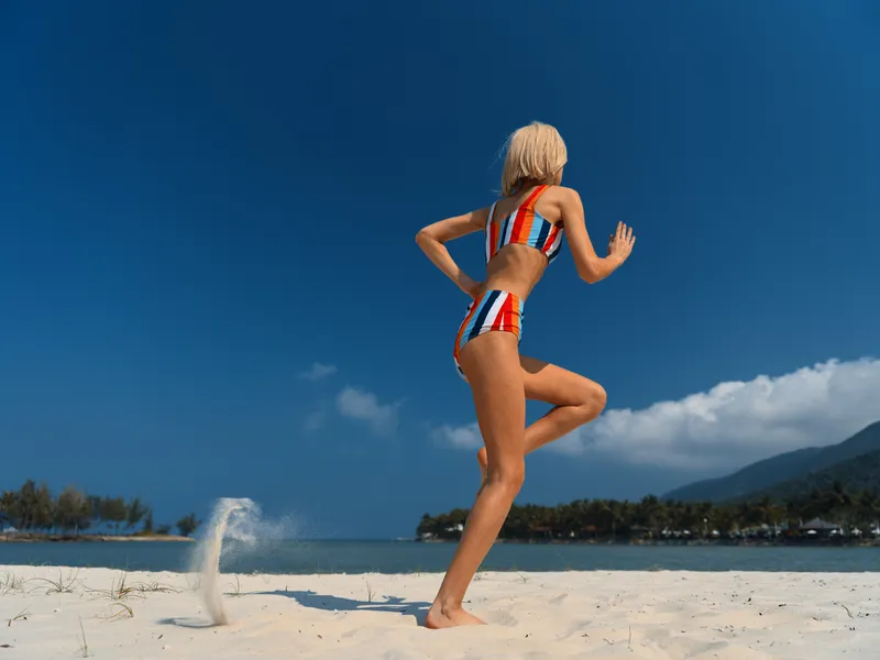 A athletic woman in a striped bikini jumps on bright beach sand beneath a clear blue sky, as water spray arcs and captures a playful moment of summer energy.