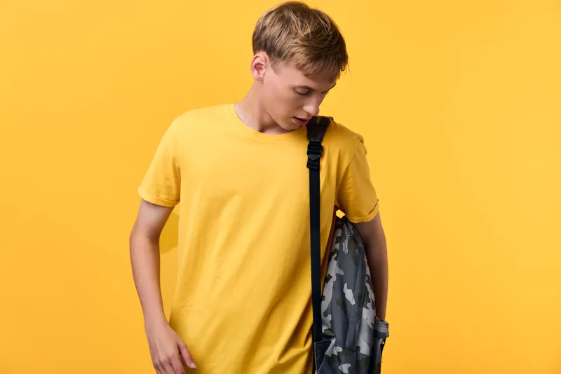 Boy in a yellow t shirt with a camouflage backpack stands against a bright yellow backdrop, casual youth style, ready for school or daily adventure