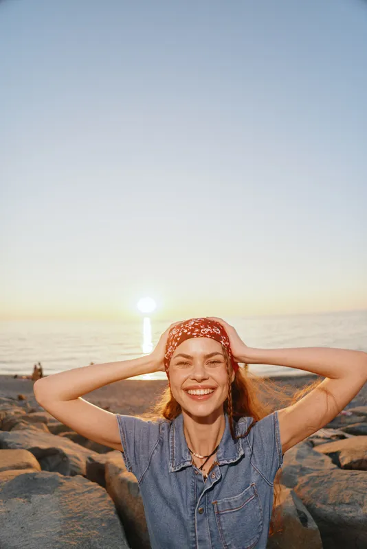 Lifestyle woman in modern boho-western denim street style enjoys sunset at the rocky beach, smiling with relaxed and joyful mood, film color warmth captured.