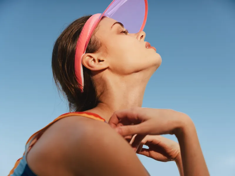Portrait of a woman wearing a pink visor, looking upward against a clear blue sky, emphasizing sunlit skin, graceful neck, and sporty summer vibe.