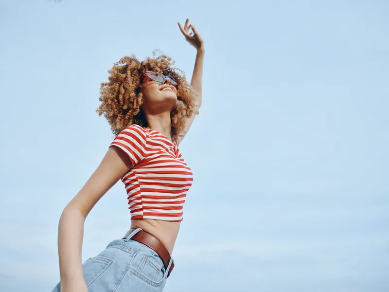 Young woman with curly hair, striped crop top and denim jeans stands against a clear blue sky, arms raised in joyful freedom and sunny confidence. Her stance exudes energy and carefree fashion.