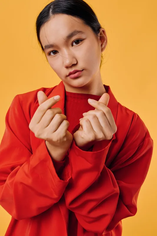 Young woman in red jacket shows finger heart gesture with serious expression on yellow background. Portrait of confident female making love sign for affection or friendship.