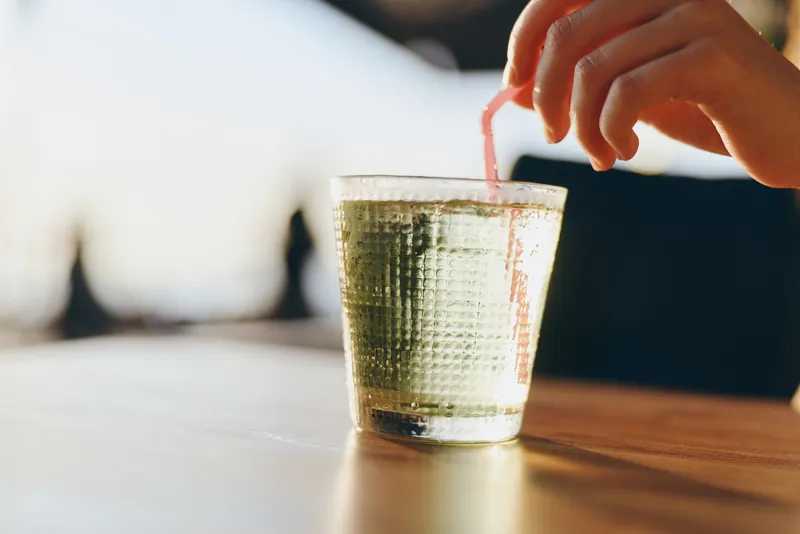 Lifestyle woman in modern boho-western denim street style stirring a drink with a straw, captured in warm film color tones, showcasing casual and trendy summer vibe.