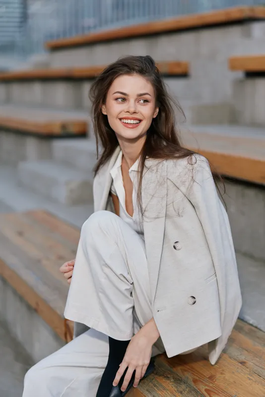 Smiling woman in light beige suit sitting on wooden stairs outdoors, cheerful young Caucasian female with natural makeup and casual hairstyle in modern fashion style