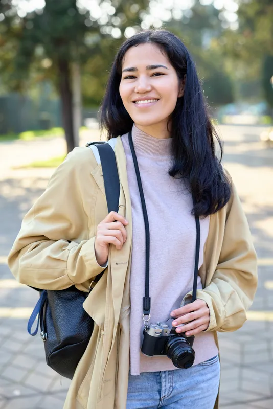 Woman with a camera outdoors in a casual jacket and light sweater, carrying a backpack, smiling as she walks along a sunlit park path, ready for street photography and travel.