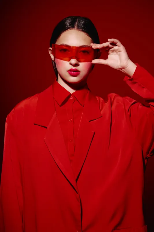 Young woman wearing oversized red blazer and transparent red visor effect glasses poses confidently with red lips styled hair and matching vibrant background and apparel.