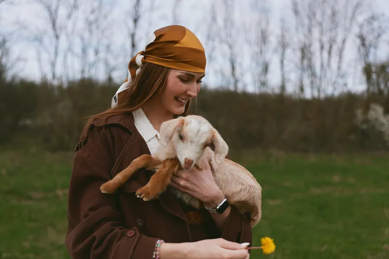 Joyful woman outdoors, cuddling a goat kid in a warm rustic setting, soft light, gentle breeze, natural tones, relaxed pose, affectionate moment with animal, countryside atmosphere