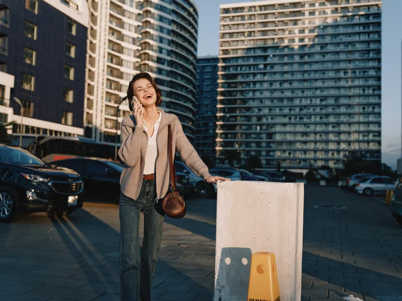 A cheerful businesswoman talks on her phone outside in a sunny parking area, with modern high rise buildings behind, wearing smart casual attire.