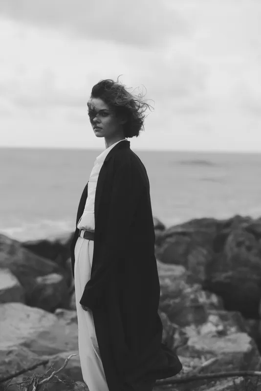 Coast, shore, wind, coat, sea, blackandwhite model gazes toward horizon in moody photography, rocks in foreground, ocean backdrop, dramatic atmosphere, minimalist styling, monochrome scene, weathered