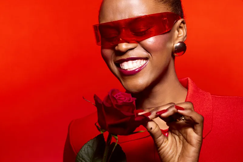 Smiling black woman with dark skin wearing red sunglasses and red outfit holds red rose on red background. Concept of fashion, beauty, confidence, and elegance in studio lighting.