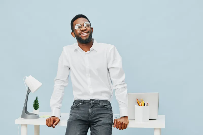 Smiling man with glasses standing confidently in office environment, casual white shirt, gray jeans, cheerful expression, modern workspace, bright background, positive mood, lifestyle concept, adultho