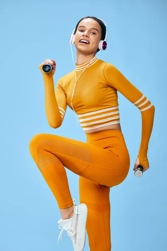 Energetic woman in bright orange fitnesswear performs a high-energy workout in a studio, lifting a dumbbell and swinging a jump rope with headphones for focused athletic training.