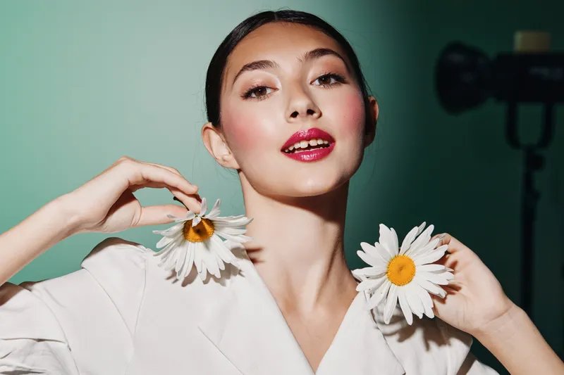 Young woman with glossy red lips holds two white daisies near her neck. Fashion makeup look with stylish nails on a dark green background. Creative beauty portrait concept.