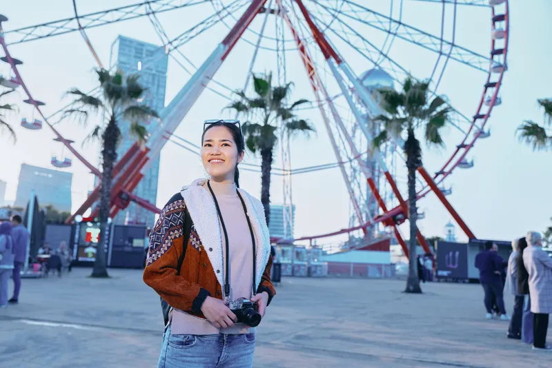 Ferris wheel amusement park scene with a smiling woman holding a camera, outdoor fashion, palm trees in the background, evening lights, candid travel photography and exploration