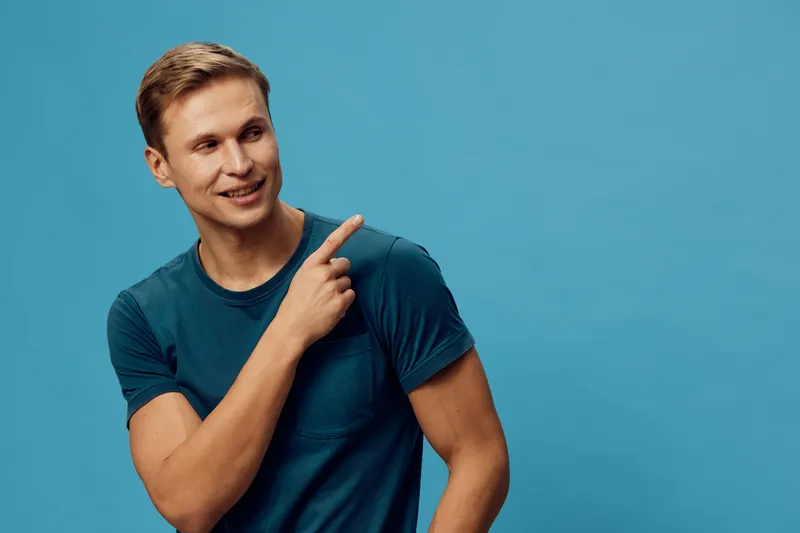 Smiling young caucasian man wear blue t-shirt point finger look side isolated on plain blue background casual style digital nomad concept