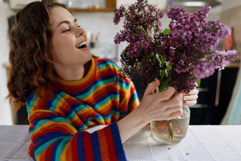Smiling woman with rainbow striped sweater admiring a vase of lilac flowers on a dining table in a cozy home setting. Warm, natural light and a cheerful, everyday lifestyle mood.