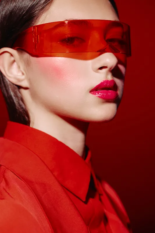 Closeup portrait of stylish woman with glossy red lips and artistic makeup wearing transparent red futuristic sunglasses on red background. Lem Portrait beauty fashion creative beauty theme.