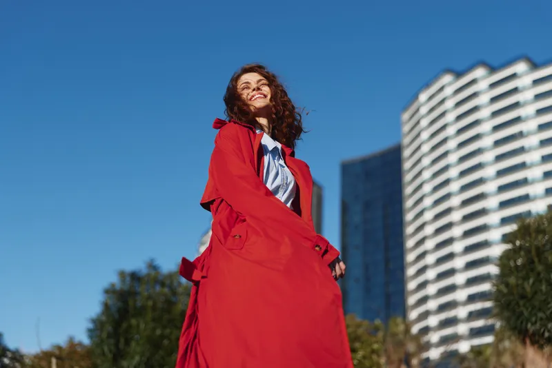 Young woman with curly hair wearing a red coat smiles confidently outdoors on a sunny day with modern buildings in the background and blue sky above.