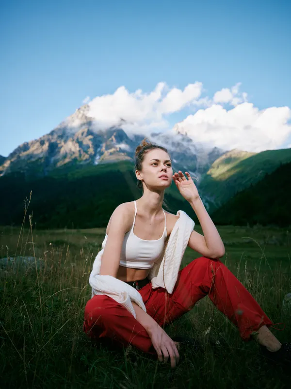 Woman sitting on a grassy field with a towel draped over her shoulder, casual white top and red pants, framed by a mountain landscape under a bright blue sky.