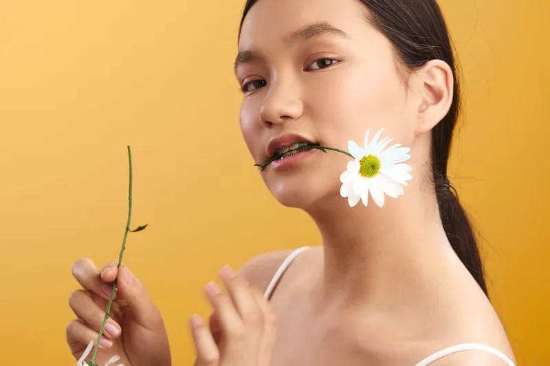 asian woman with a peaceful expression holds a daisy near her lips in a bright studio, creating a fresh, natural mood that conveys elegance and serenity for lifestyle and fashion use.
