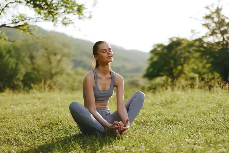 Young woman practicing yoga in nature, enjoying tranquility, wearing comfortable sportswear, surrounded by greenery and sunlight, promoting mindfulness and well being