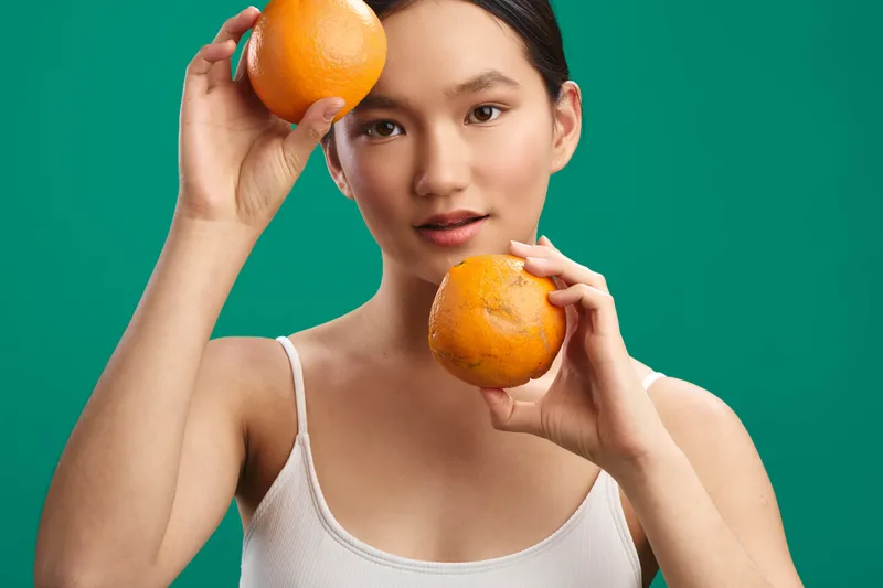 asian woman holds oranges against a vivid green background, radiating energy and freshness as she playfully frames the fruit near her face in a bright studio portrait.