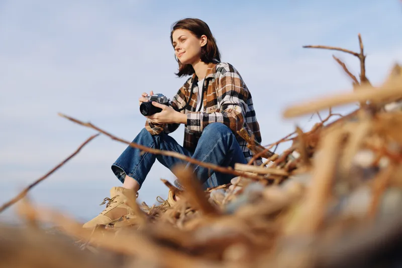 Photographer woman sitting on driftwood, holding a camera in casual plaid shirt and jeans, exploring outdoors with daylight light, focused on gear and natural landscape