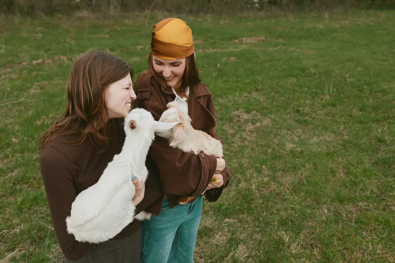 Two young women enjoy a casual outdoor moment holding and feeding a baby goat in a grassy field, sharing smiles and connection in a relaxed lifestyle setting.