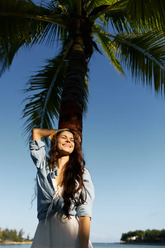 Smiling woman standing under palm tree wearing light summer dress and denim jacket with clear blue sky background, warm sunlight, and tropical atmosphere.