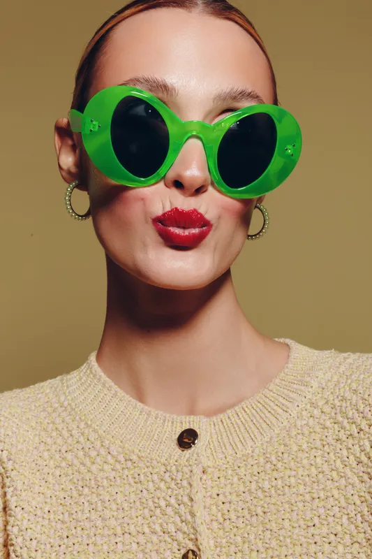 Young woman with bright green sunglasses and red lipstick makes a playful kiss face. She wears a beige knitted sweater and gold hoop earrings on a neutral background.
