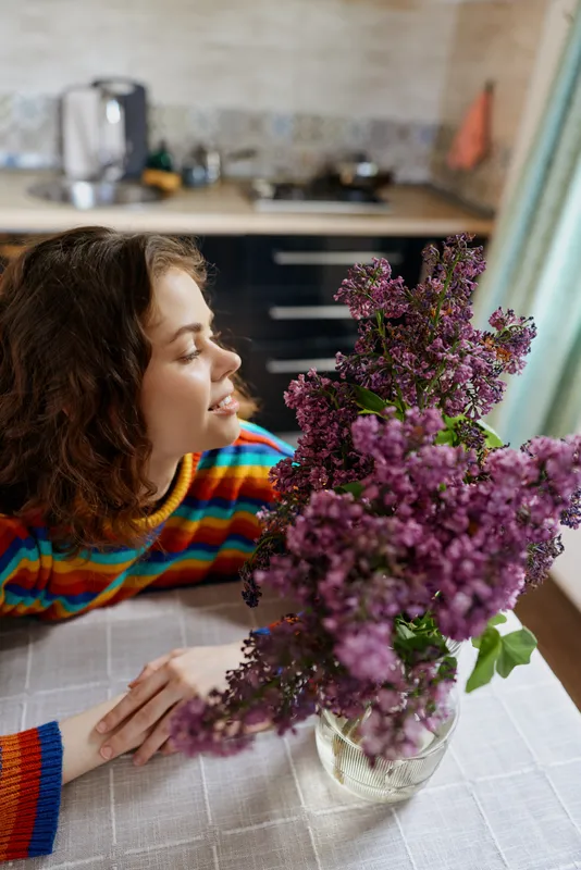 A young woman indoors leans toward a vase of purple lilacs on a kitchen table, smiling softly in warm natural light, enjoying a calm cozy moment of everyday joy.