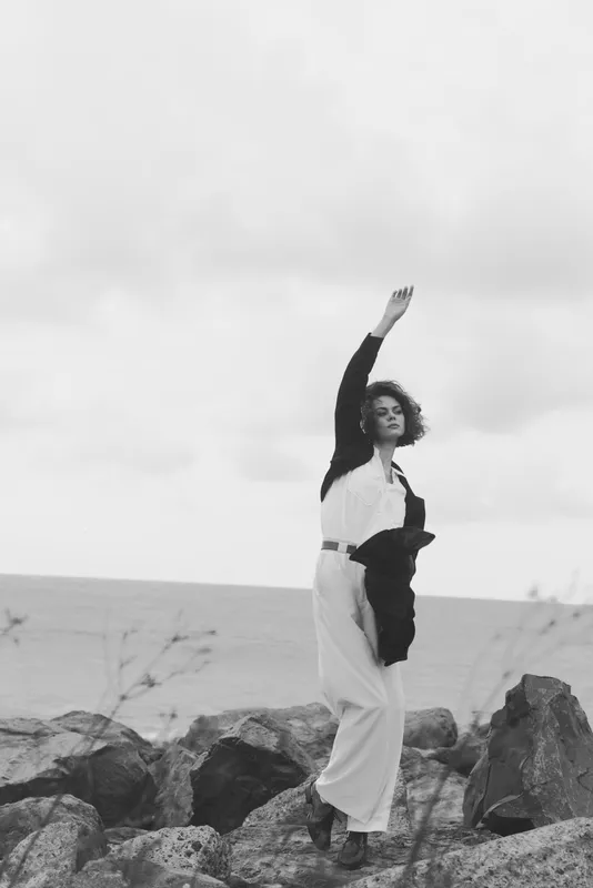 Woman on rocky shore in monochrome dress strikes a bold pose as wind lifts hair, ocean backdrop, outdoor scene, fashion and freedom