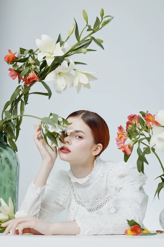 Elegant female model surrounded by fresh flowers, showcasing a stylish white outfit and soft makeup, conveying grace and tranquility in a spring inspired concept
