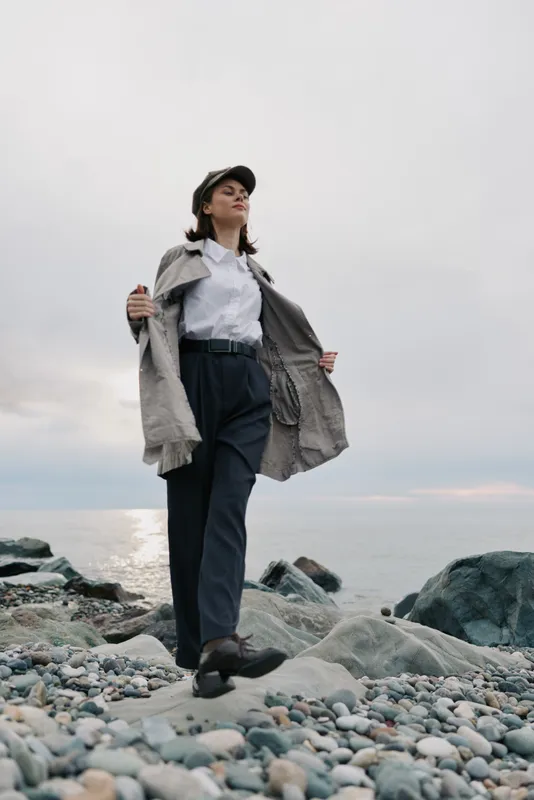 Confident young woman in casual clothes and cap walks on rocky beach near calm sea under cloudy sky. Concept of freedom, fashion, and outdoor lifestyle in nature.