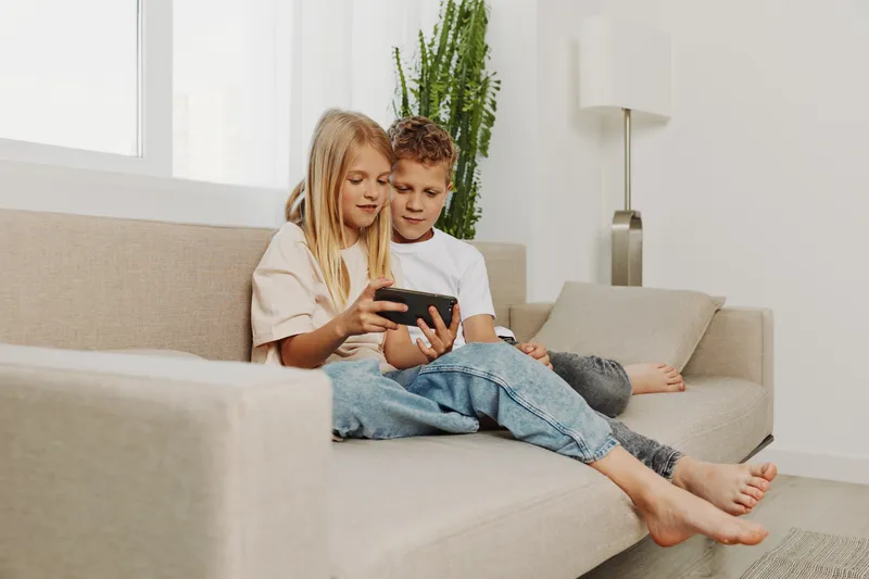 Two children sitting closely on a beige sofa playing with a smartphone, showing concentration and engagement. Bright living room with natural light creates cozy and relaxed atmosphere for family