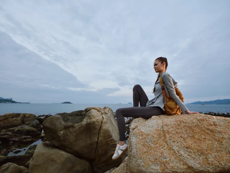 Young woman sitting on a rock by the sea, wearing casual outdoor clothing, focused on the horizon, conveying a sense of tranquility and adventure in nature