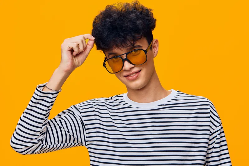 Young man with curly hair wearing yellow sunglasses and striped shirt smiles confidently against vibrant orange background, showcasing casual style and cool attitude.