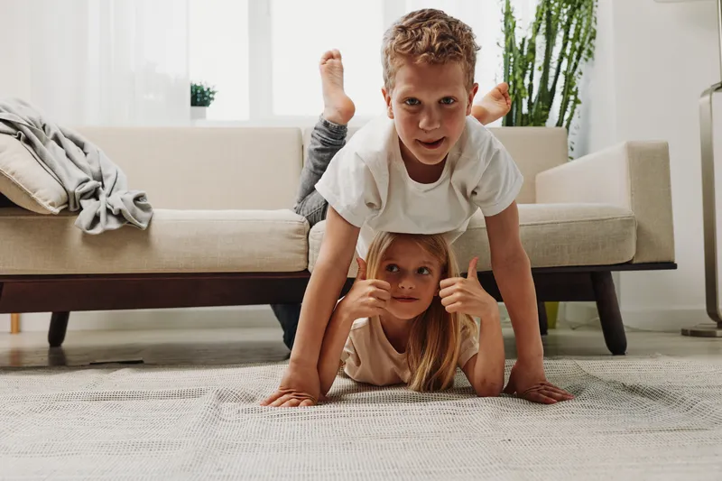 Two playful children in casual clothes enjoy a fun moment indoors on a carpet near a sofa and plants. The boy balances on the girl’s back creating a joyful family atmosphere.