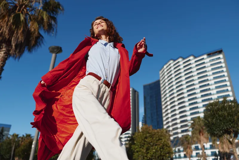 A heroic young woman in a red cape strides along an urban street, posing confidently under a bright blue sky with modern buildings behind