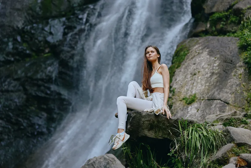 Young woman sitting on rock near waterfall in forest, wearing white activewear and sneakers, enjoying nature and outdoor adventure, peaceful and relaxed atmosphere.