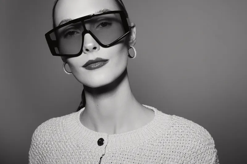 Stylish white woman with pale skin wearing oversized sunglasses and hoop earrings poses in knitted sweater. Minimalist black and white portrait with high fashion concept and confident mood.