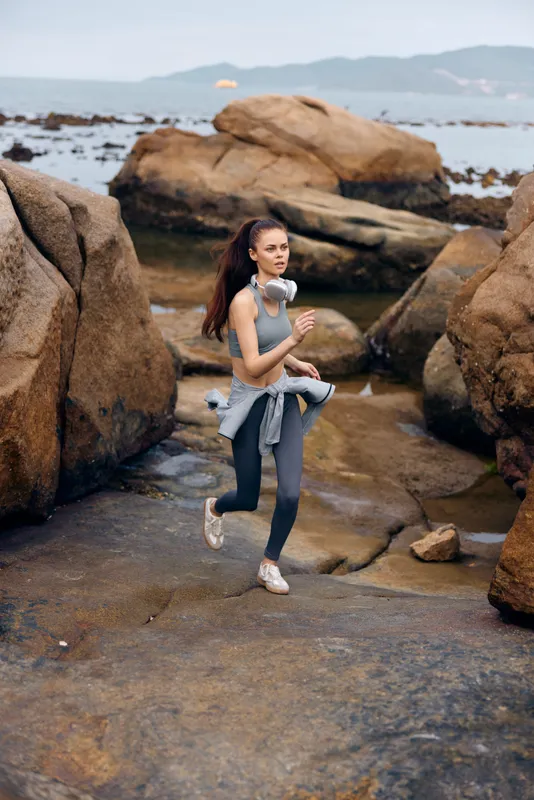 Young woman jogging on rocky beach in activewear, showcasing fitness and determination, with calming ocean background and natural elements, ideal for health concepts